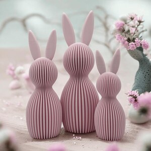 Può includere: Tre figurine di coniglio rosa con una texture a coste. I conigli sono di dimensioni diverse e sono disposti in fila.