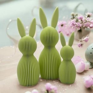 Può includere: Tre figurine di coniglio di colore verde e con una texture a coste. I conigli sono di dimensioni diverse e sono disposti in fila su una superficie rosa chiaro.