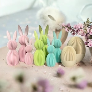 Può includere: Un set di sette decorazioni di coniglietti di Pasqua color pastello. I coniglietti sono fatti di carta e hanno un design plissettato. Ci sono tre coniglietti rosa, due coniglietti verdi e due coniglietti blu. I coniglietti sono disposti in fila su una superficie marrone chiaro.