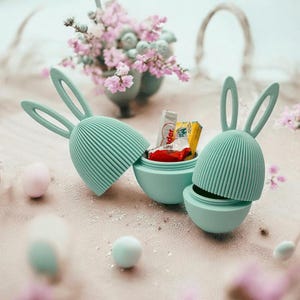 Puede incluir: Dos contenedores de huevos de Pascua en forma de conejo de color verde pastel con un pequeño paquete de caramelos dentro de uno de los contenedores. Los contenedores están abiertos y los caramelos son visibles.