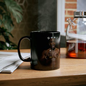 Puede incluir: Taza de café de cerámica negra con un diseño que cambia de color. La taza presenta una foto de una persona con el texto "BOO!" impreso en la taza.