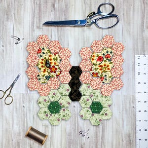May include: A handmade butterfly quilt block composed of colorful fabric hexagons. The wings feature floral patterns in red, cream, and green. Sewing scissors, pins, a ruler, and thread are arranged around the quilt block on a wood-grain surface.