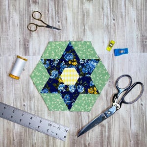 May include: A patchwork star with blue floral fabric and a yellow diamond centre. The star is surrounded by green floral fabric. Sewing tools, including scissors, a ruler, thread, and clips, are arranged around the star on a wooden surface.
