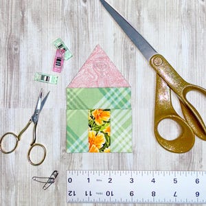 Puede incluir: Un bloque de casa de tela verde y rosa con un patrón floral, una regla, tijeras y pequeñas pinzas sobre una superficie de madera.