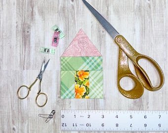 Op de afbeelding: Een groen en roze stoffen huisblok met een bloemenpatroon, een liniaal, een schaar en kleine clips op een houten oppervlak.