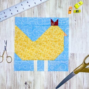 May include: A square fabric block featuring a yellow chicken design on a blue background with white stripes. The chicken has a red accent. Sewing tools, including scissors, clips, and a ruler, are scattered around the block on a wooden surface.