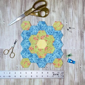 May include: A patchwork quilt block made of blue, green, and yellow hexagonal fabric pieces. The block is surrounded by gold scissors, pins, and clips. A ruler is placed at the bottom, measuring up to 30 cm.