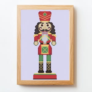 May include: A framed cross-stitch of a traditional nutcracker soldier. The nutcracker is depicted in red, green, and gold, with a black mustache and hair. The background is a light purple color.