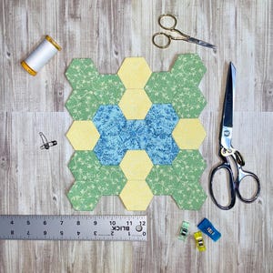 May include: A patchwork hexagon quilt block in yellow, green, and blue floral patterns. Sewing tools include scissors, thread, a ruler, and clips. The ruler measures up to 12 inches.