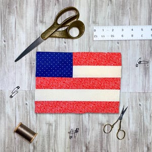 May include: A fabric American flag with red and white stripes and a blue field with white dots. Gold scissors, a ruler, safety pins, and a spool of thread are arranged around the flag on a wood-grain surface.