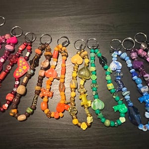 May include: Ten colorful beaded keychains with silver key rings. The keychains are made with different colored beads and charms, including hearts, stars, and other shapes. The keychains are arranged in a rainbow pattern, with the colors red, orange, yellow, green, blue, indigo, and violet.