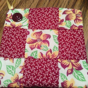 May include: A red and white quilted hot pad with a floral pattern and a brown button. The hot pad is made of fabric and is perfect for protecting surfaces from heat.