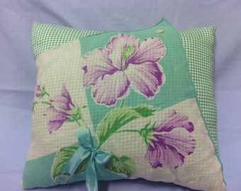 Puede incluir: Cojín cuadrado con un diseño floral con flores de hibisco moradas sobre un fondo azul claro y blanco. El cojín tiene un estampado de cuadros verdes en los laterales y una cinta azul claro atada con un lazo.