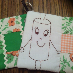 May include: A white fabric pouch with a hand-stitched design of a smiling stick figure. The pouch is made of patchwork fabric with green, white, and orange floral patterns.