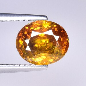 May include: An oval-cut gemstone with a vibrant orange and golden yellow color palette. The stone features a faceted surface that reflects light, creating a sparkling appearance with internal patterns of deep amber and bright yellow hues.