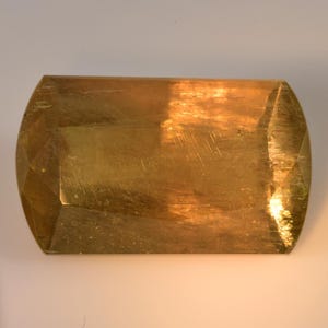 May include: A rectangular, gold-colored object with rounded ends and faceted edges. The surface has a brushed texture and reflects light, creating highlights and shadows. The overall shape resembles a large gemstone.