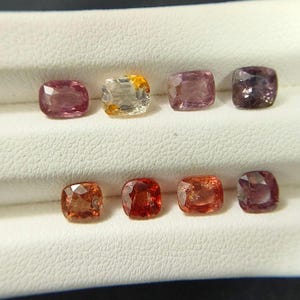 May include: Eight cushion-cut gemstones arranged in two rows. The top row features a pink, a clear with yellow, a light purple, and a dark purple stone. The bottom row displays three orange stones of varying intensity and one dark purple stone.