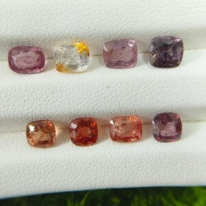 May include: Eight cushion-cut gemstones arranged in two rows on a white display stand. The stones feature a variety of colors including shades of pink, orange, yellow, and deep purple, each with a faceted surface that reflects light.
