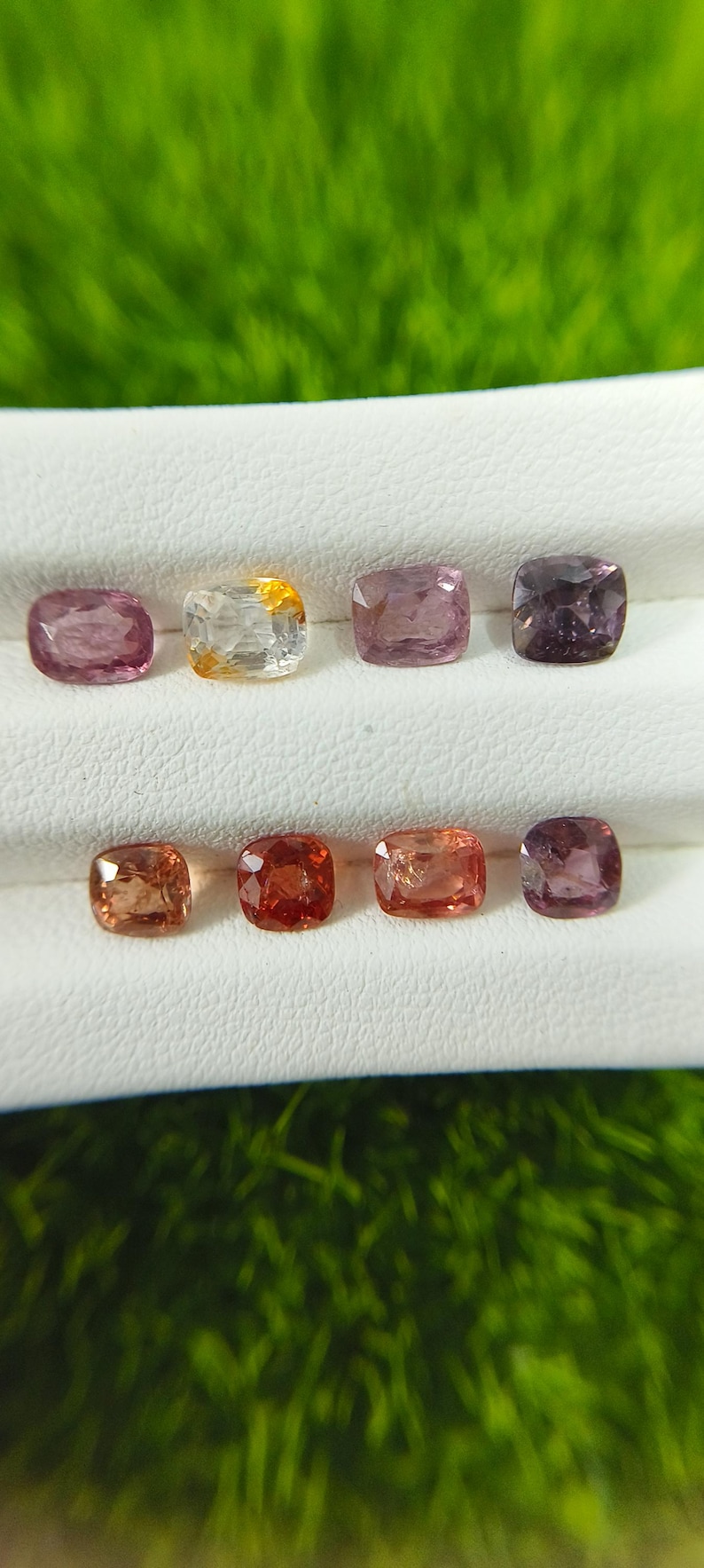 May include: Eight cushion-cut gemstones arranged in two rows on a white display stand. The stones feature a variety of colors including shades of pink, orange, yellow, and deep purple, each with a faceted surface that reflects light.
