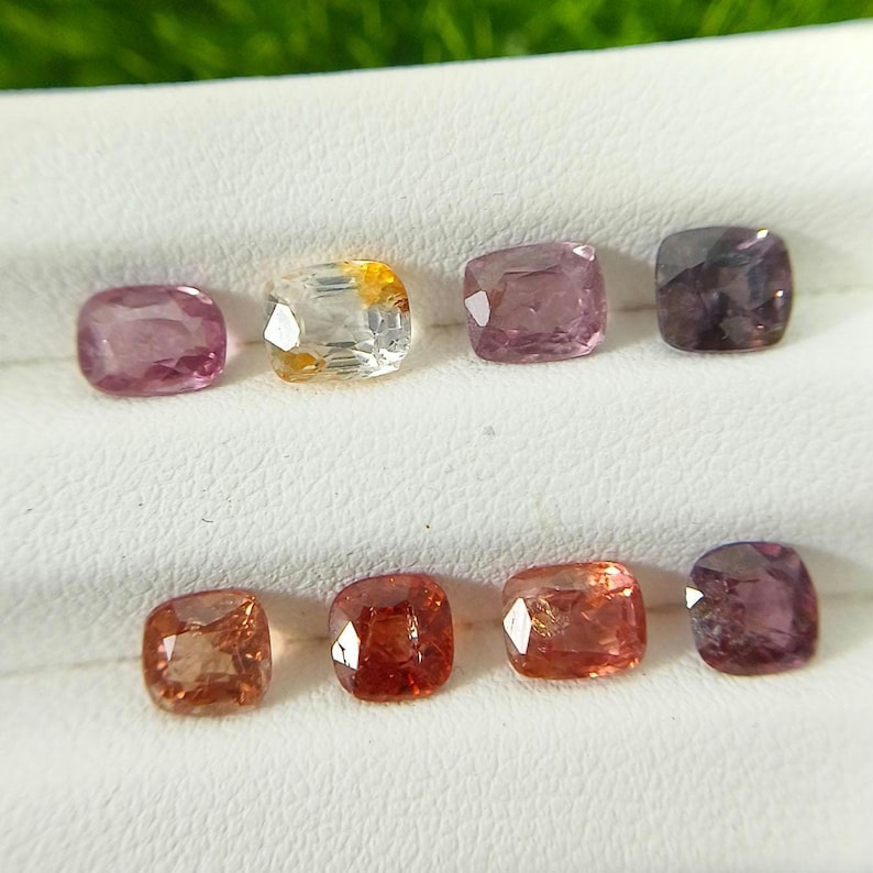 May include: A collection of eight faceted cushion-cut gemstones arranged in two rows. The stones display a variety of colors including shades of pink, purple, orange, and a clear stone with yellow inclusions, all featuring a polished, reflective surface.