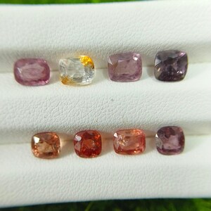 May include: Eight cushion-cut gemstones arranged in two rows. The top row features a light pink stone, a white and yellow stone, a mauve stone, and a dark purple stone. The bottom row displays a light orange, a deep red, a peach, and a plum stone.