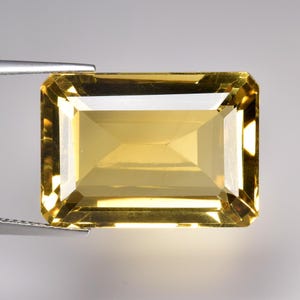 May include: A radiant, rectangular-cut gemstone with a golden-yellow colour. The gem is held by silver tweezers, highlighting its precise facets and polished surface. The gemstone's cut creates a play of light, enhancing its sparkle.
