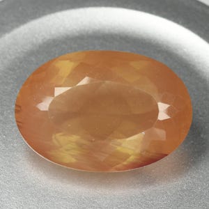 May include: An oval-shaped, faceted gemstone with warm orange and yellow hues. The gem has a polished surface with multiple facets that catch the light, creating a sparkling effect. The gemstone is set against a neutral background.