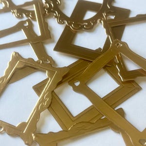 May include: A collection of flat, gold-colored metal frames in various rectangular and oval shapes. Each frame features distinct border details, including smooth edges and ornate, scalloped patterns. The frames are arranged in an overlapping pile.