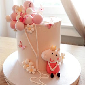Könnte beinhalten: Ein hellrosa Kuchen, verziert mit rosa Kugeln, weißen und gelben Blumen und rosa Schmetterlingen. Der Kuchen zeigt eine Peppa Wutz-Figur mit Krone und der Zahl 3. Der Kuchen steht auf einer weißen Kuchenplatte.
