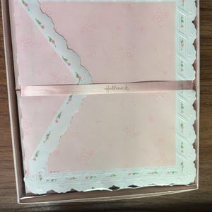 May include: A boxed set of light pink stationery featuring a delicate white scalloped border with small pink floral accents. The paper is secured with a light pink ribbon printed with the Hallmark brand name in gold script lettering.