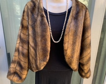 May include: A brown and black faux fur bolero jacket with a rounded collar and long sleeves. The jacket is displayed on a mannequin with a black dress and a pearl necklace. The jacket's fur has a striped pattern.