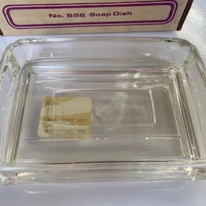 May include: A clear, rectangular glass soap dish with a slightly raised rim. The dish has a rectangular base with a series of parallel lines. The top of the box reads "No. 556 Soap Dish". A small sticker is visible inside the dish.