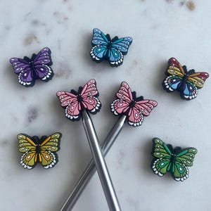 May include: A selection of butterfly charms in shades of purple, blue, pink, yellow, and green, with white detailing. The charms are arranged around two silver knitting needles. The background is a white marble surface.