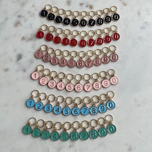 May include: A selection of stitch markers, each with a number from 0 to 9. The markers are small, circular charms in various colours, including black, red, pink, and teal. Each charm has a gold-coloured ring.