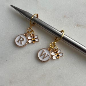 May include: Gold-toned stitch markers with white enamel circles featuring the letters "R" and "W", and small bee charms. The stitch markers are hanging on a silver knitting needle. The bees have black and yellow stripes.