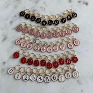 May include: Several rows of number charms in various colors: black, pink, red, and white. Each charm features a number from 0 to 9 in a contrasting color, set on a gold-colored chain. The charms are arranged on a light-colored surface.