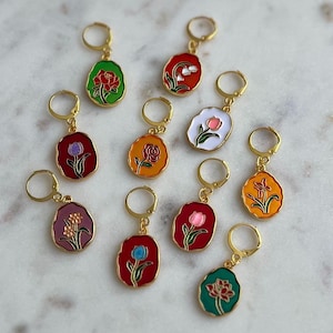 Peut inclure: Une collection de boucles d'oreilles créoles dorées. Chaque boucle d'oreille présente un pendentif ovale coloré avec un motif floral. Les fleurs sont représentées dans diverses couleurs, dont le rouge, le rose, l'orange et le violet, avec des feuilles vertes.