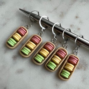 May include: Five keyrings with miniature macarons in pink, yellow, and green. Each keyring has a rectangular, tan-coloured base. The macarons are attached to silver-coloured key rings.