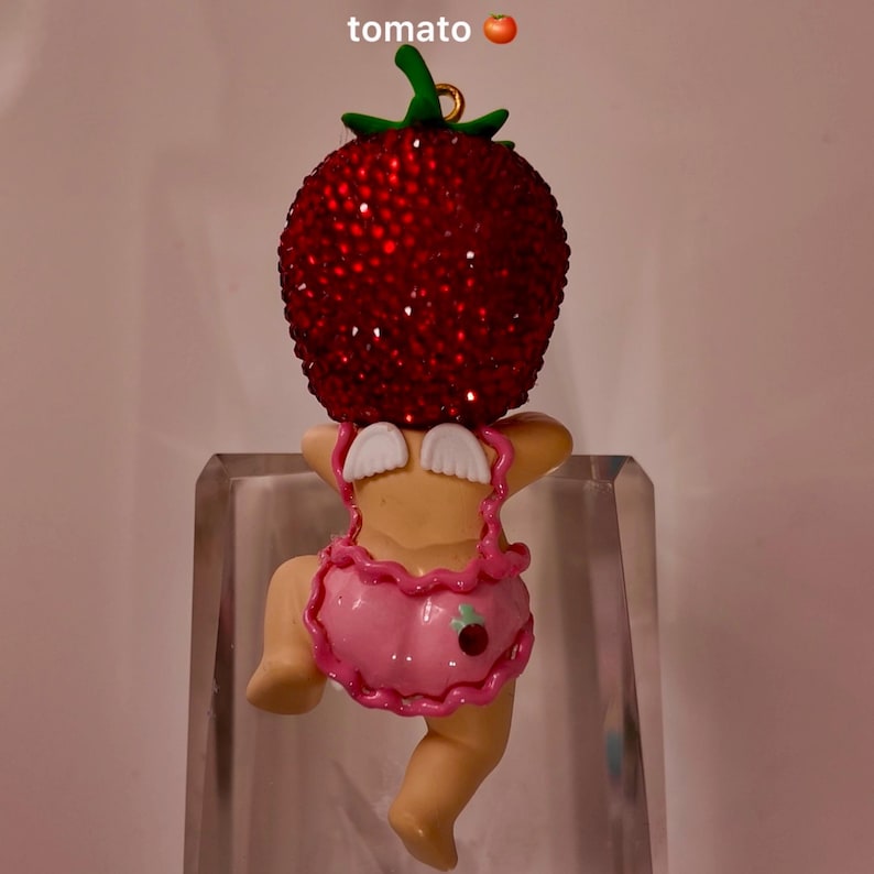 Puede incluir: Una figura caprichosa con una cabeza de tomate roja con incrustaciones de joyas sobre un cuerpo de querub&iacute;n. La figura lleva un atuendo rosa con un detalle de cereza. La palabra "tomato" est&aacute; escrita encima de la figura.