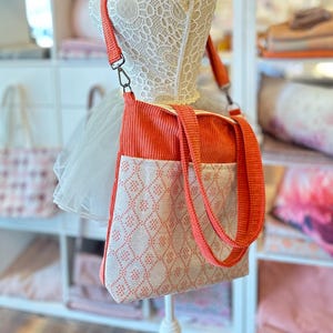May include: A coral-colored tote bag with a white and pink geometric pattern. The bag has two long straps and a shorter strap for carrying on the shoulder.