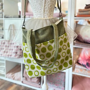May include: A green and white tote bag with a polka dot pattern. The bag has two top handles and a long shoulder strap.
