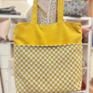 May include: A yellow tote bag with a checkered pattern on the bottom half. The bag has two thick yellow straps.