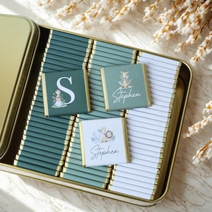May include: An open gold-colored tin filled with individually wrapped chocolates. Some chocolates have a green background with a white letter "S" or the name "Stephen" with animal illustrations. The chocolates are arranged on a green and white striped background.
