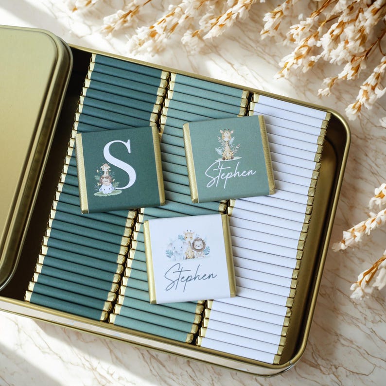 May include: An open gold-colored tin filled with individually wrapped chocolates. Some chocolates have a green background with a white letter "S" or the name "Stephen" with animal illustrations. The chocolates are arranged on a green and white striped background.