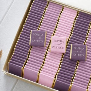 Pu&ograve; includere: Una scatola riempita con file di barrette di cioccolato rettangolari avvolte in carta color viola e rosa con bordi in foglia d'oro. Tre barrette recano la scritta bianca Sophia and Patrick in un carattere tipografico serif.