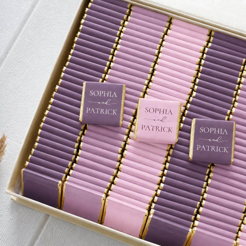 Pu&ograve; includere: Una scatola riempita con file di barrette di cioccolato rettangolari avvolte in carta color viola e rosa con bordi in foglia d'oro. Tre barrette recano la scritta bianca Sophia and Patrick in un carattere tipografico serif.