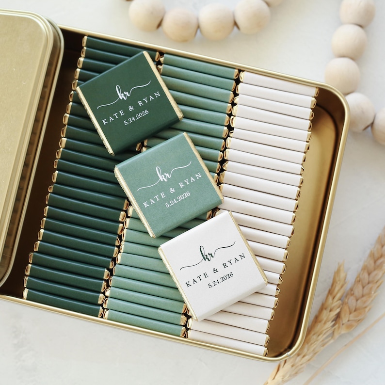 Pu&ograve; includere: Una scatola di metallo dorato riempita con file di piccole barrette di cioccolato rettangolari avvolte in carta verde scuro, verde salvia e bianca. Tre barrette presentano il testo stampato kr, KATE & RYAN e 5.24.2026 in carattere script e serif.