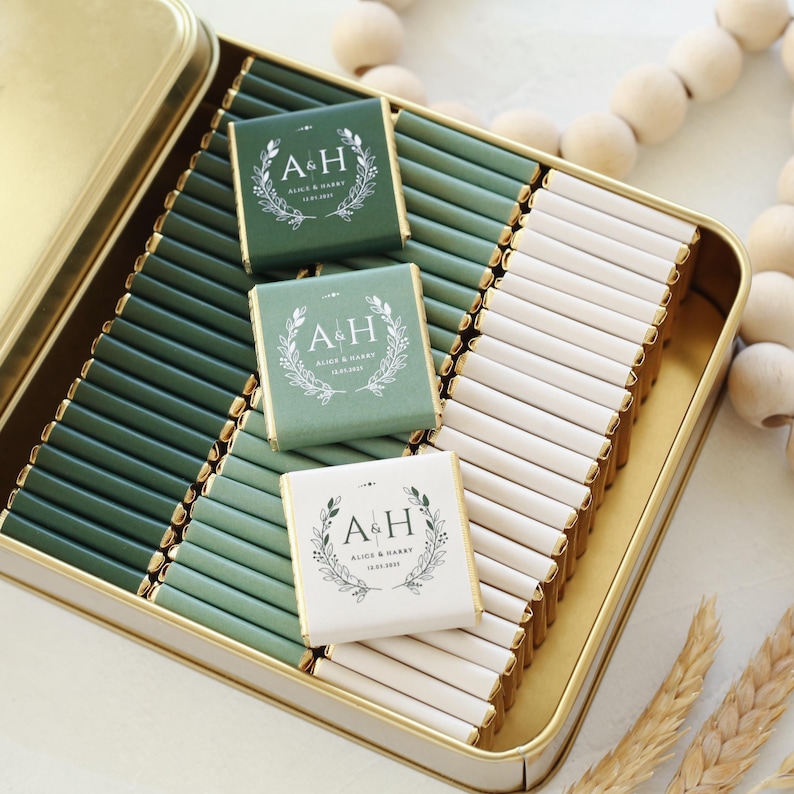 Pu&ograve; includere: Una scatola di metallo color oro riempita con file di piccole barrette di cioccolato rettangolari avvolte in carta verde scuro, verde salvia e bianca. Tre barrette presentano un disegno di corona d'alloro bianca con le lettere A & H.