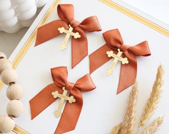 May include: Three rust-coloured satin bows with gold cross charms, each bearing the name "Sophia." The bows are arranged on a white surface with gold trim, alongside wheat stalks and wooden beads. A decorative and elegant presentation.