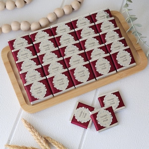 May include: A wooden tray holds numerous small chocolate bars with burgundy ribbon and silver foil. Each bar features a decorative label with the names "YAJAIRA and TYLER". The chocolates are arranged neatly, with a few loose bars nearby.